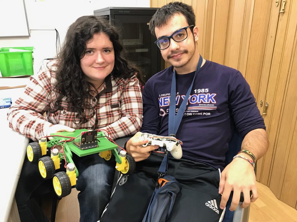 Silvia y Javier mantienen en sus manos el coche rover y el mando de XBOX modificado para que funcionen juntos. El coche, impreso en 3D, tiene seis ruedas y es verde y amarillo.
